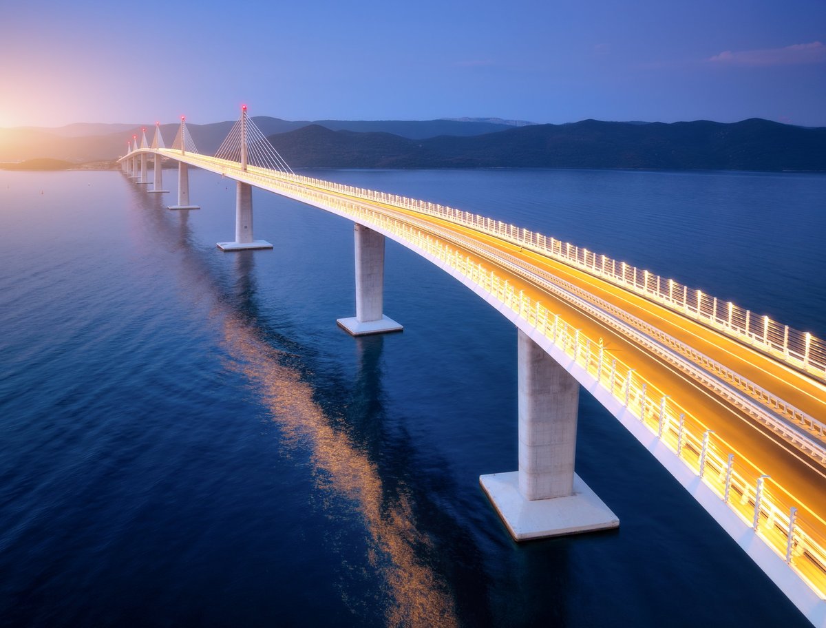 Illuminated bridge crossing toward mountains at golden-hour sunset
